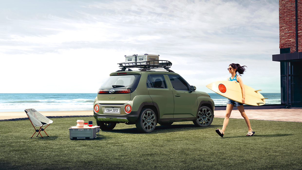 잔디밭 위를 걸어가는 서핑보드를 든 여성과 현대자동차 캐스퍼의 후측면 모습