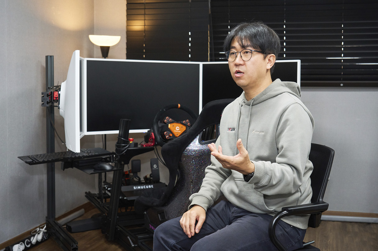 레이싱 시뮬레이터를 배경으로 남성이 의자에 앉아 설명하는 모습