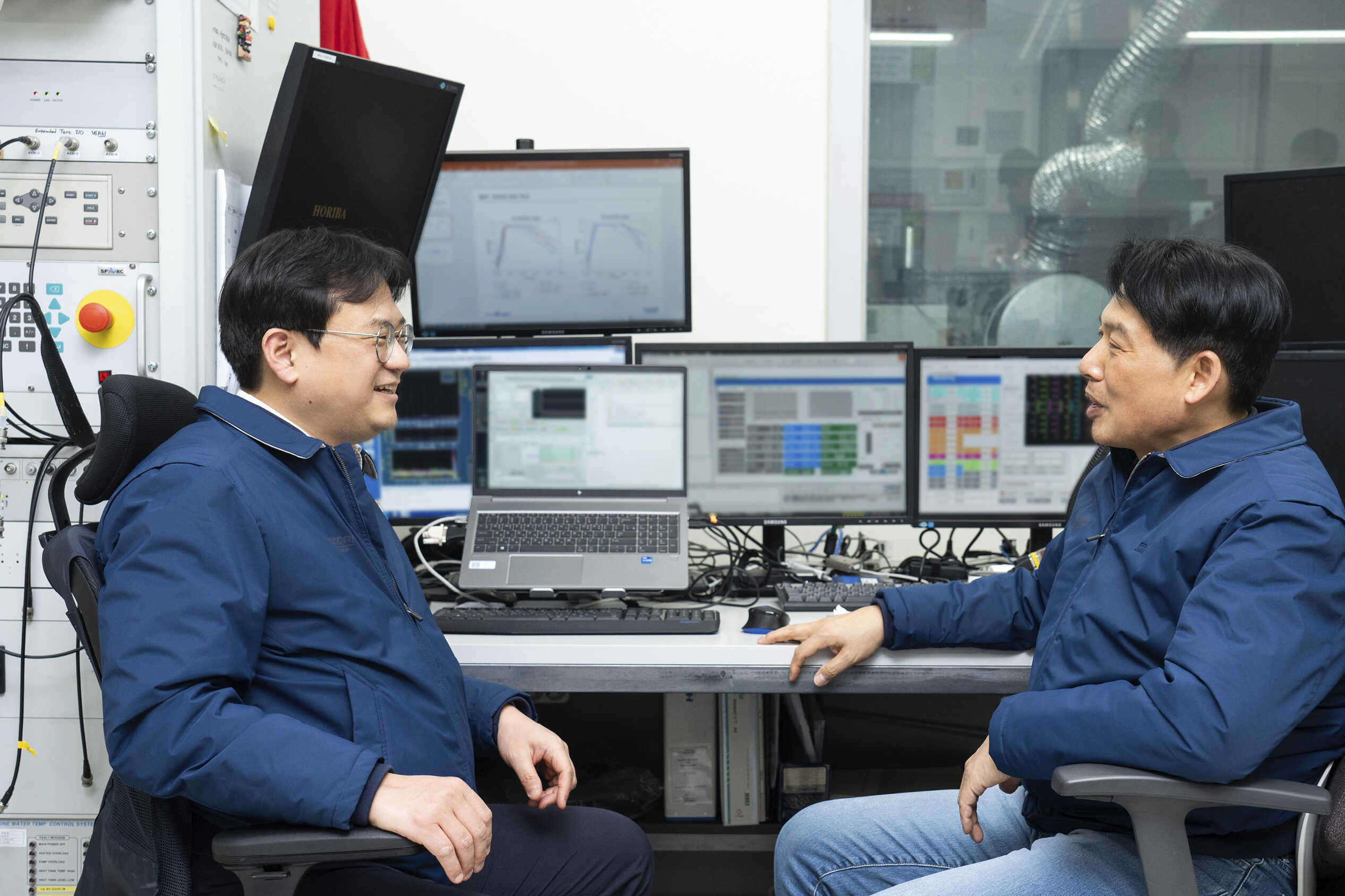 성인 남성 두 명이 파란색 점퍼를 입은 채 의자에 앉아 서로 마주보고 대화하는 모습