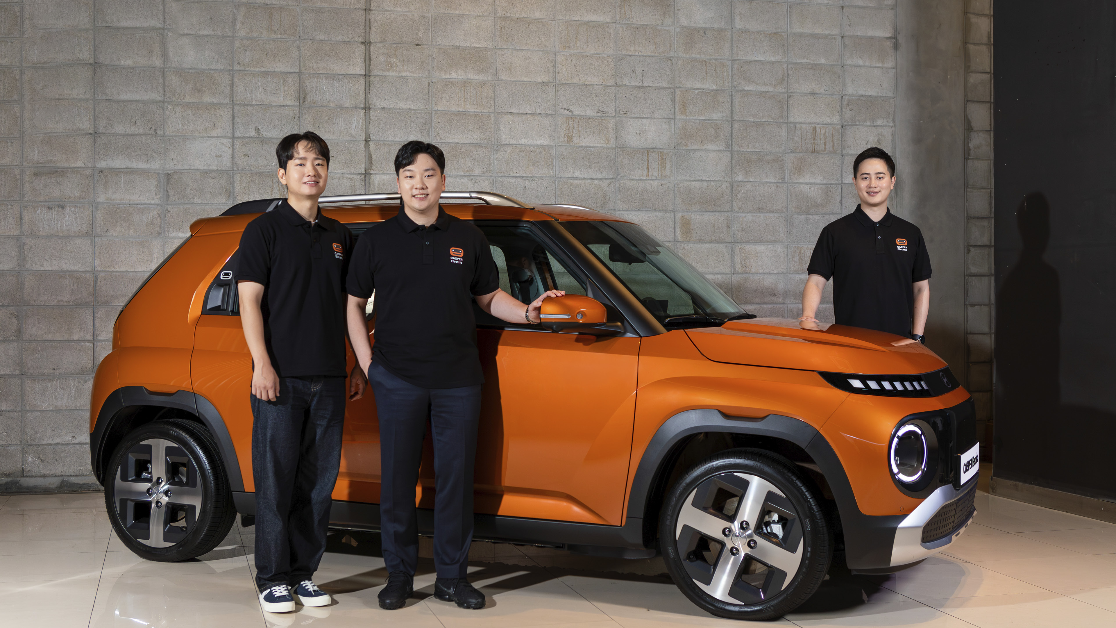 오렌지 경차 앞에 성인 남자 세 명이 서 있는 모습
